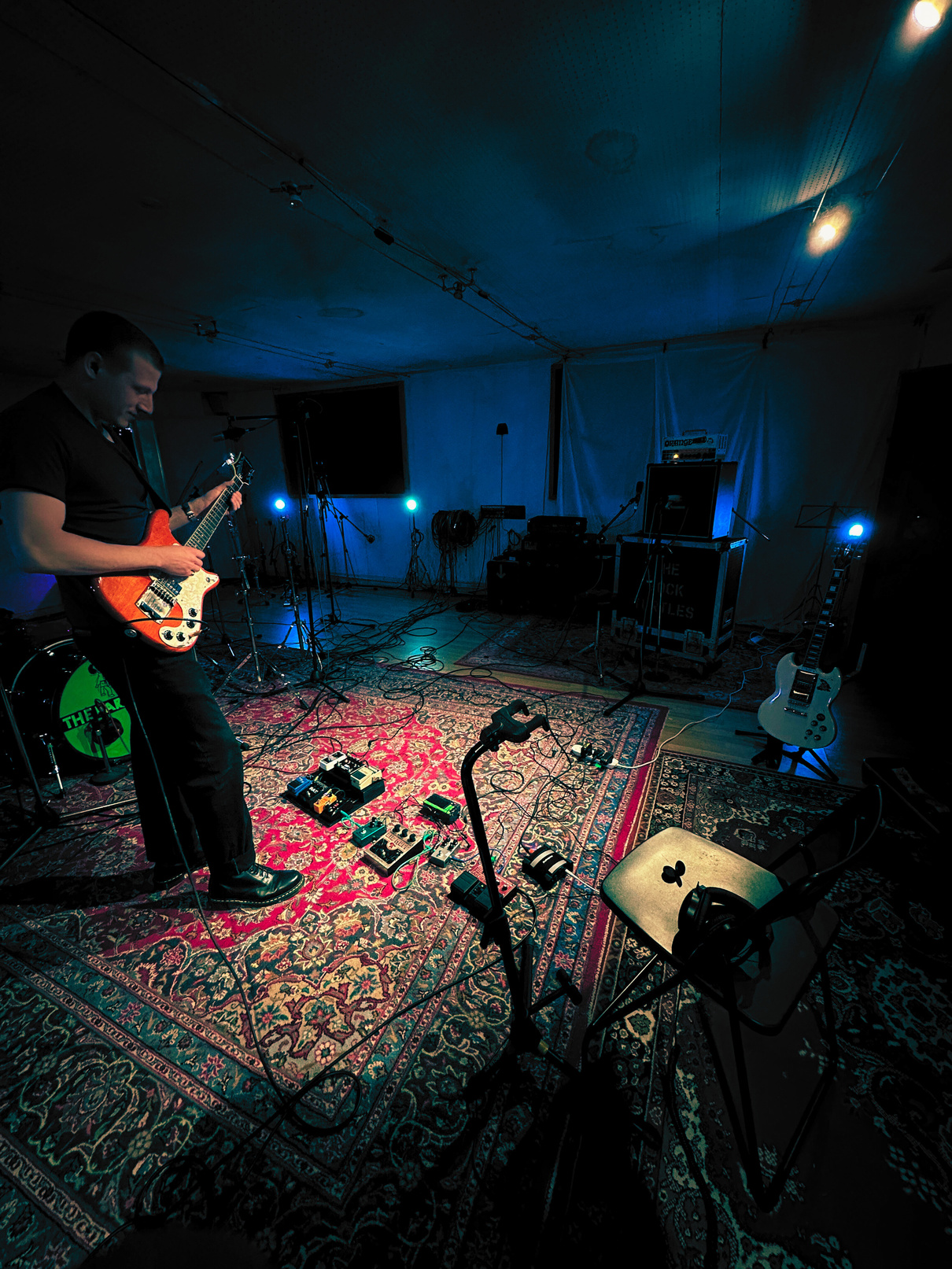 Boni playing guitar in the live room, staring down at his pedals set over a backdrop of toned down blue lighting with the fancy rug underneath everything.