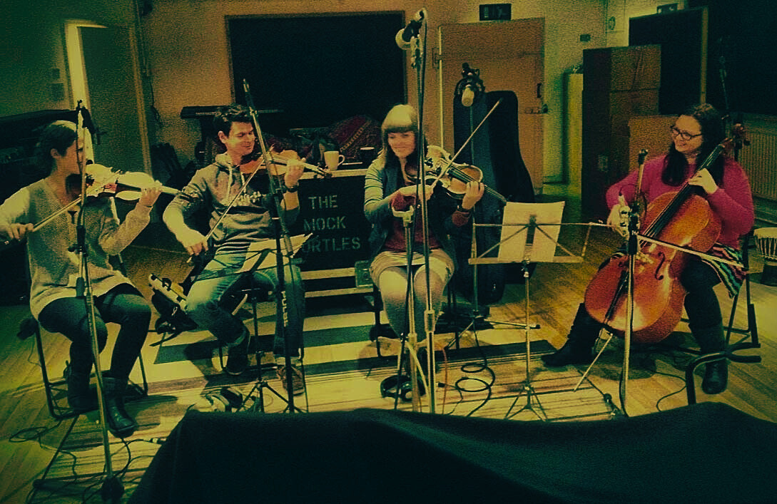 a string quartet play three violins and a cello between them, all smiling and having a good time sat in a semi circle in the middle of the live room at VIBE Recording Studio, Manchester